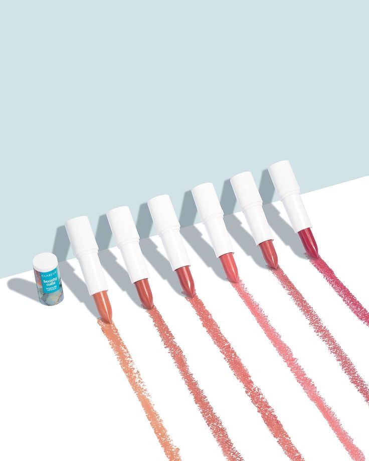 Kreatywny packshot: sześć szminek w białych opakowaniach, każda w innym odcieniu, pozostawia ślad koloru na białym tle z błękitnym akcentem. Obok jedna szminka w opakowaniu.