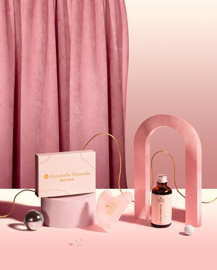 Fotografia produktowa Annabelle Minerals: pudełko Gua Sha, kamień Gua Sha i butelka Stay Essential na tle różowych rekwizytów i drapowanej tkaniny.