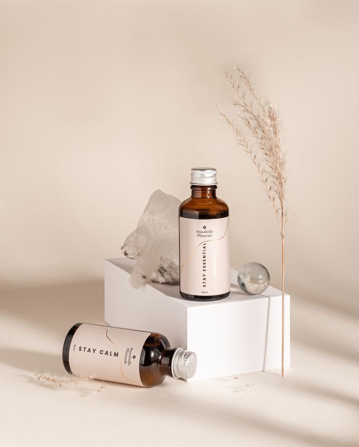 Dwie butelki Annabelle Minerals na białym postumencie z kryształem i szklaną kulką, obok źdźbło trawy. Jedna butelka leży, druga stoi. Stonowana kolorystyka, minimalistyczny styl.