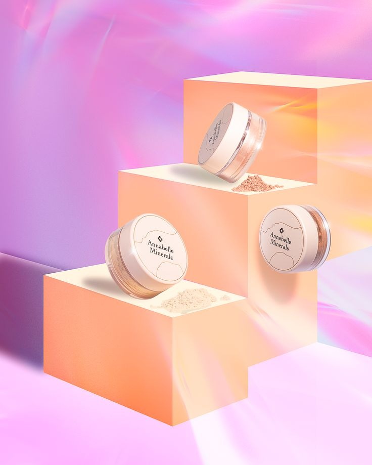 Zdjęcie produktowe: trzy słoiczki z pudrem Annabelle Minerals na geometrycznych platformach w pastelowych kolorach. Profesjonalne oświetlenie podkreśla detale.