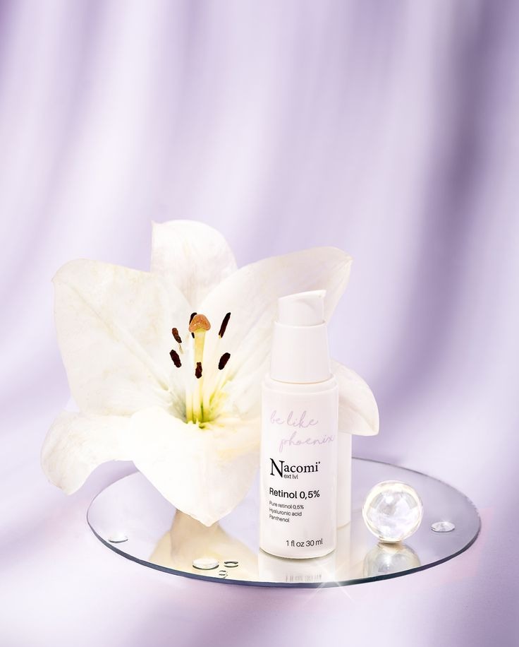 Stylizowany packshot serum Nacomi Retinol 0,5% na lustrzanej podstawce z lilią i szklaną kulką na tle delikatnej fioletowej tkaniny. Elegancka kompozycja.