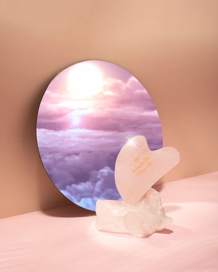 Różowy kamień Gua Sha Annabelle Minerals na krysztale, odbicie nieba w okrągłym lustrze, pastelowe tło. Zdjęcie produktowe.