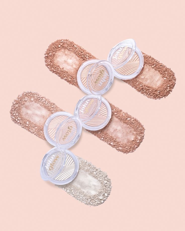 Stylizowana fotografia produktowa: trzy pudry rozświetlające John Glow, otwarte, ułożone na smugach rozsypanego pudru na jasnoróżowym tle.