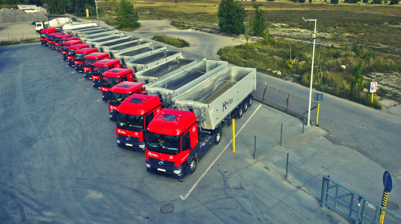 Widok z góry na rząd czerwonych ciężarówek z naczepami wypełnionymi materiałem sypkim, zaparkowanych na placu; w tle widoczny krajobraz polny.