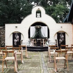 Uroczysta aranżacja przestrzeni na zewnątrz: rzędy drewnianych krzeseł z kremowymi wstążkami, ozdobiona kwiatami scena z białymi zasłonami, łapacze snów na drzwiach, białe lampiony, kamienna ściana...