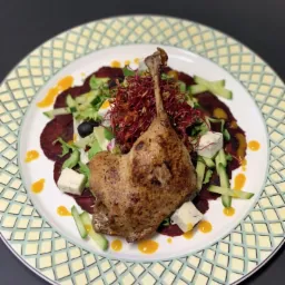 Pieczona noga kaczki na carpaccio z buraka z dodatkiem ogórka, sera pleśniowego, sałaty i czerwonych kiełków, polana sosem na talerzu ze wzorem geometrycznym.