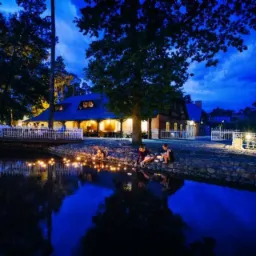 Oświetlony budynek restauracji z tarasem nad wodą, wieczorny plener. Trzy osoby siedzą nad brzegiem, w wodzie pływają lampiony.