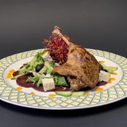 Wykwintne danie: pieczona noga kaczki podana na carpaccio z buraka z sałatką, serem pleśniowym, ogórkiem i sosem pomarańczowym na ozdobnym talerzu.