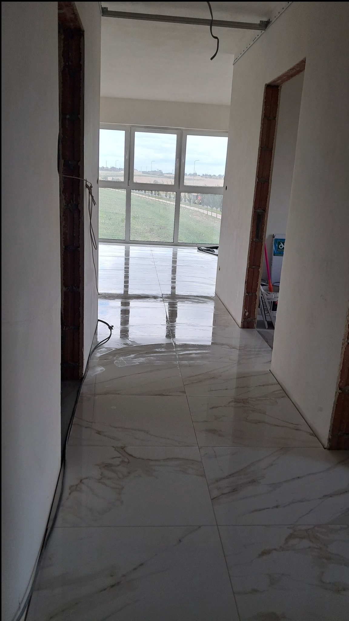 Połyskująca podłoga z płytek imitujących marmur w korytarzu w trakcie remontu. Widoczne niedokończone ściany i okno z widokiem na zewnątrz.