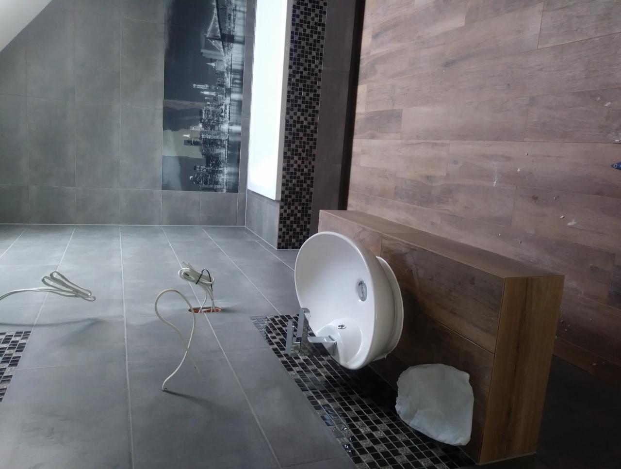 Niedokończona łazienka z szarymi płytkami podłogowymi i ściennymi, drewnopodobną ścianą, podwieszaną umywalką i wystającymi przewodami elektrycznymi.