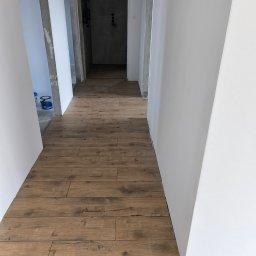 fliesen-tech - Korytarz po remoncie: brązowe panele podłogowe imitujące drewno, białe ściany, surowy beton przy wejściu. Widok w głąb pomieszczenia.