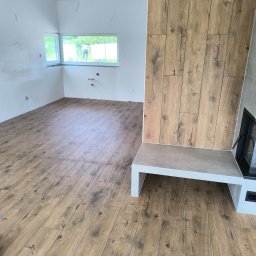 fliesen-tech - Drewnopodobne panele podłogowe w jasnym pokoju z kominkiem i oknem, przez które widać osobę na zewnątrz.