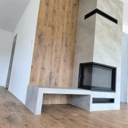 fliesen-tech - Nowoczesny kominek z obudową z płytek imitujących beton i drewno, z czarnym wkładem kominkowym. Podłoga wyłożona panelami imitującymi drewno. Minimalistyczny design.