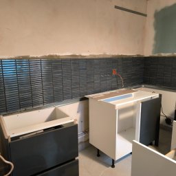 fliesen-tech - Montaż grafitowej mozaiki w kuchni, z widocznymi fragmentami instalacji elektrycznej i niedokończoną zabudową meblową, z kuchenką gazową w tle.