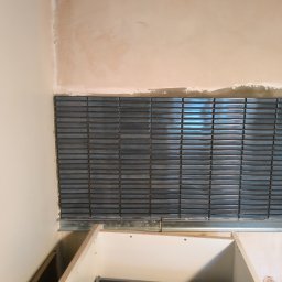 fliesen-tech - Układanie błyszczącej, podłużnej, niebieskiej mozaiki na ścianie, widoczny fragment niezabudowanej szafki. Ściana częściowo przygotowana pod dalsze prace.