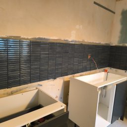 fliesen-tech - Montaż ciemnych, poziomych płytek w kuchni, widoczne częściowo zamontowane szafki kuchenne, surowa ściana powyżej płytek, gniazdko elektryczne.