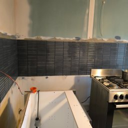 fliesen-tech - Montaż płytek mozaikowych w kuchni, widoczny fragment ściany z niebieską mozaiką, surowa ściana z płyt gipsowych i fragment kuchenki. Etap prac wykończeniowych.