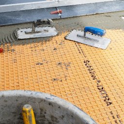 fliesen-tech - Układanie płytek na balkonie w Strzegomiu. Widoczne narzędzia glazurnicze, poziomica, wiadro z zaprawą i płytki z systemem poziomowania. Praca w toku.