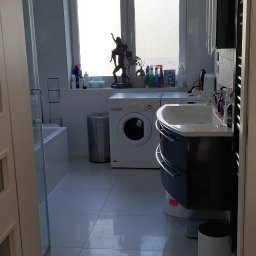 fliesen-tech - Jasna łazienka z oknem, pralką i umywalką z szafką. Na parapecie rzeźba i kosmetyki. Białe płytki podłogowe i drzwi. Widok z korytarza.