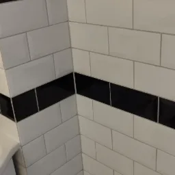 Wykończona ściana łazienki z białymi, prostokątnymi płytkami i poziomymi pasami czarnych płytek. Widoczny fragment szafki i podłogi.