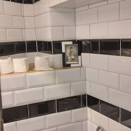 fliesen-tech - Biała toaleta z widocznymi białymi, prostokątnymi płytkami z czarnym pasem, drewnianą półką z papierem toaletowym i ramką ze zdjęciem.