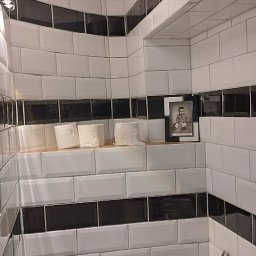 fliesen-tech - Biała toaleta w łazience z biało-czarnymi kafelkami ułożonymi w pasy. Na półce rolki papieru i czarno-białe zdjęcie. Klasyczny styl.