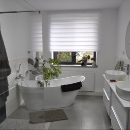 fliesen-tech - Jasna łazienka z wolnostojącą wanną, rośliną i dwoma umywalkami na blacie. Minimalistyczny design, okrągłe lustra i rolety okienne.