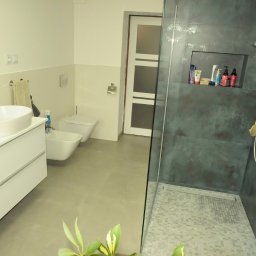fliesen-tech - Nowoczesna łazienka z kabiną typu walk-in, umywalką nablatową i podświetlanym lustrem. Ciemne płytki w strefie prysznica kontrastują z jasnymi ścianami.
