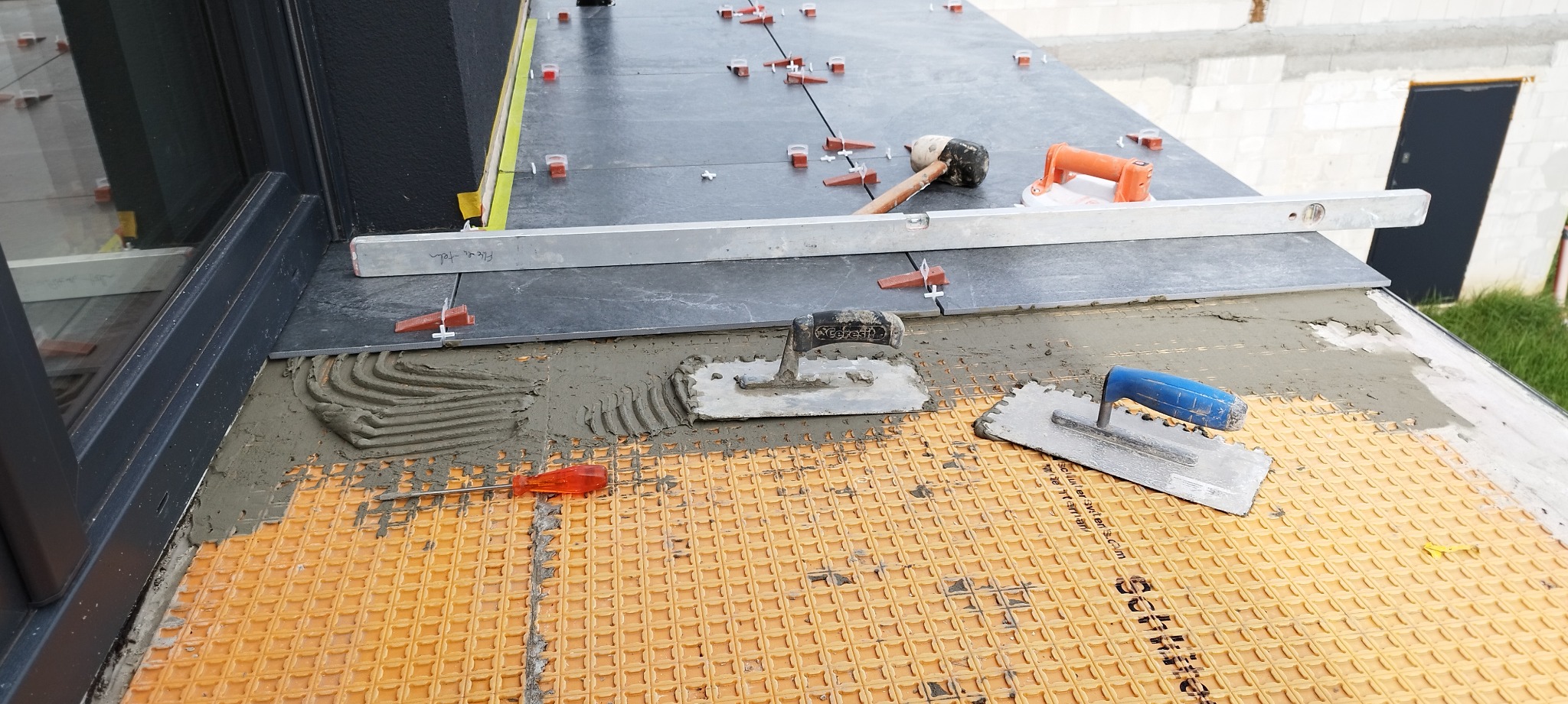 Układanie ciemnych płytek na balkonie z użyciem poziomicy, kielni i systemu poziomowania. Widoczna pomarańczowa mata drenażowa i fragment okna.