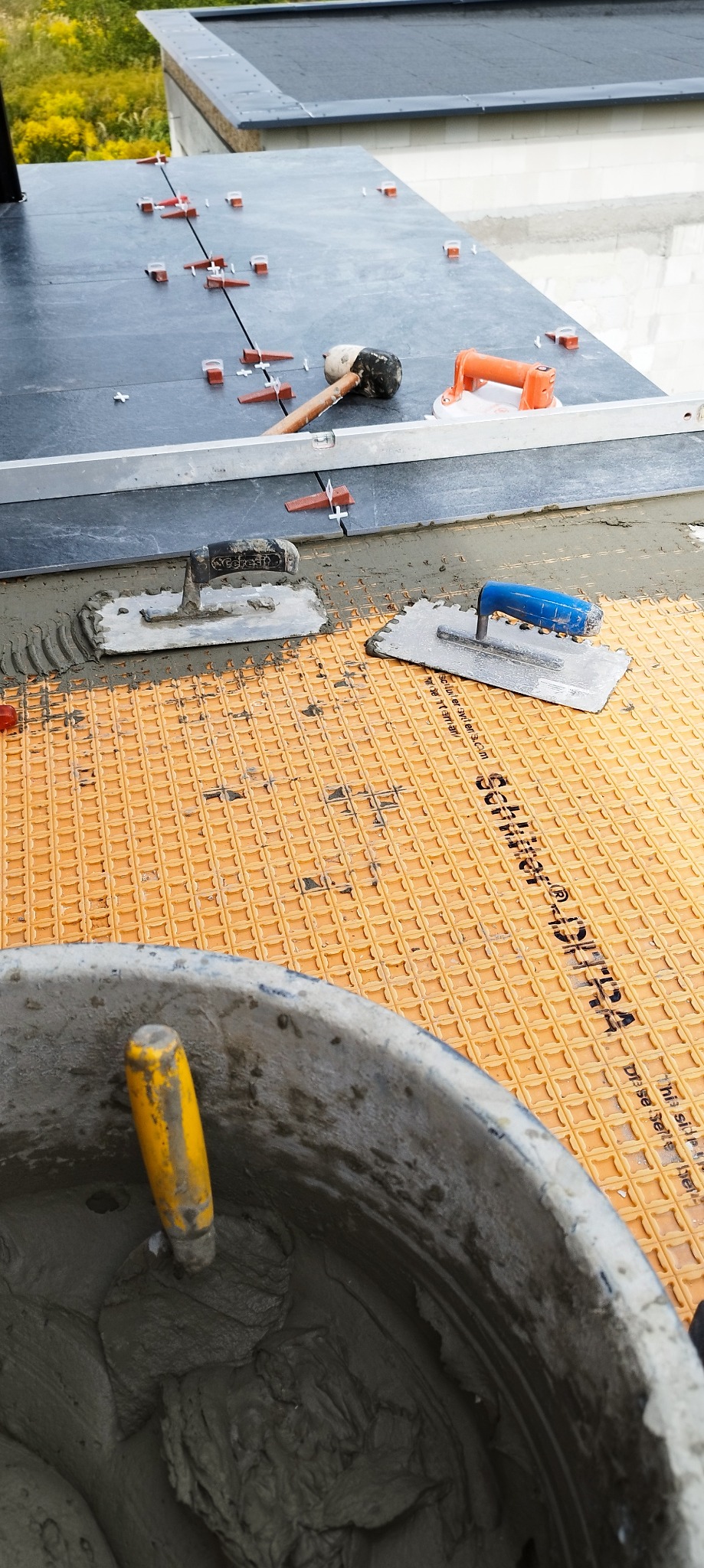 Układanie płytek na balkonie w Strzegomiu. Widoczne narzędzia glazurnicze, poziomica, wiadro z zaprawą i płytki z systemem poziomowania. Praca w toku.