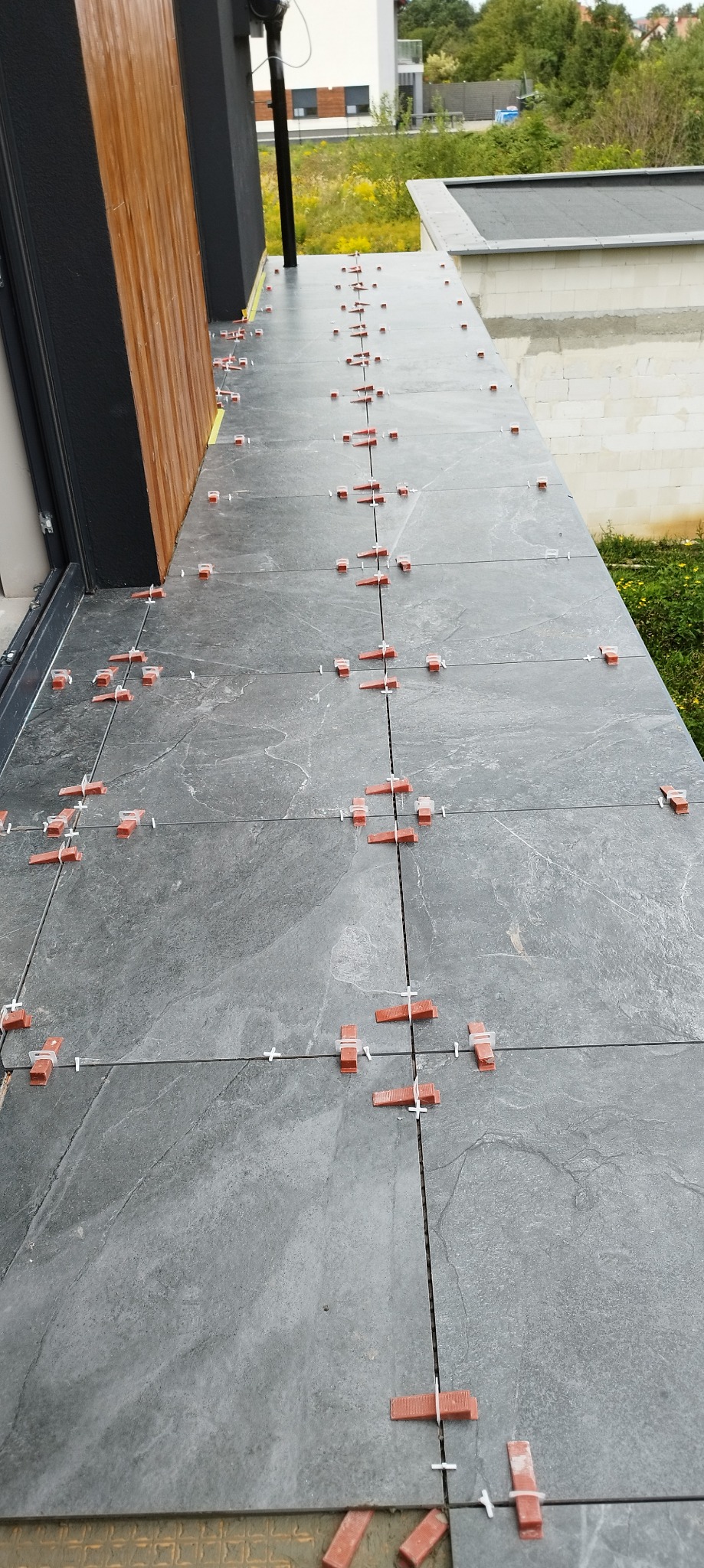 Szare płytki na balkonie w trakcie układania, z użyciem poziomujących klipsów i krzyżyków dystansowych. Widok z góry, na zewnątrz budynku, w tle zieleń.