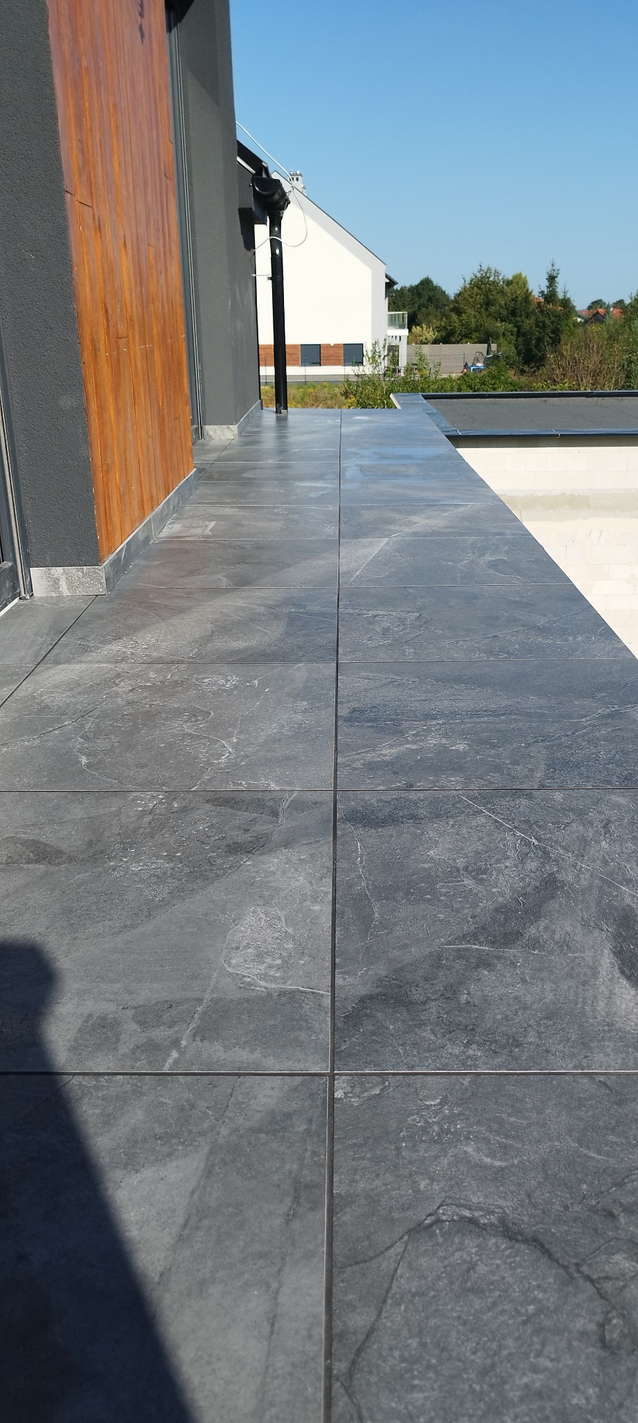 Wykończony balkon z szarymi płytkami o teksturze kamienia, widok z góry, nowoczesne wykończenie, fragment elewacji budynku i błękitne niebo w tle.