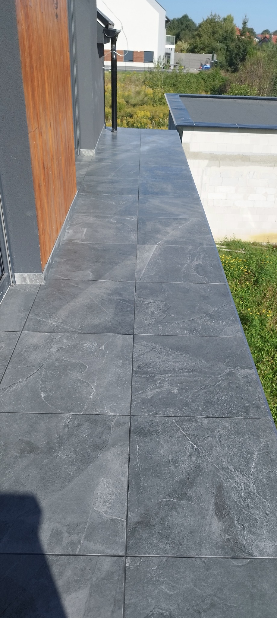 Szare płytki gresowe na balkonie, widok z góry, fuga w kolorze grafitowym. W tle widać zieleń i zabudowę jednorodzinną, słoneczny dzień.