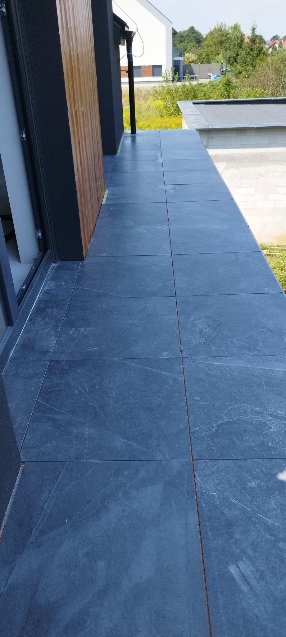 Wykończony balkon z ciemnoszarą, matową terakotą. Widoczna elewacja budynku i fragment otoczenia z zielenią. Dyskretne oświetlenie zewnętrzne.