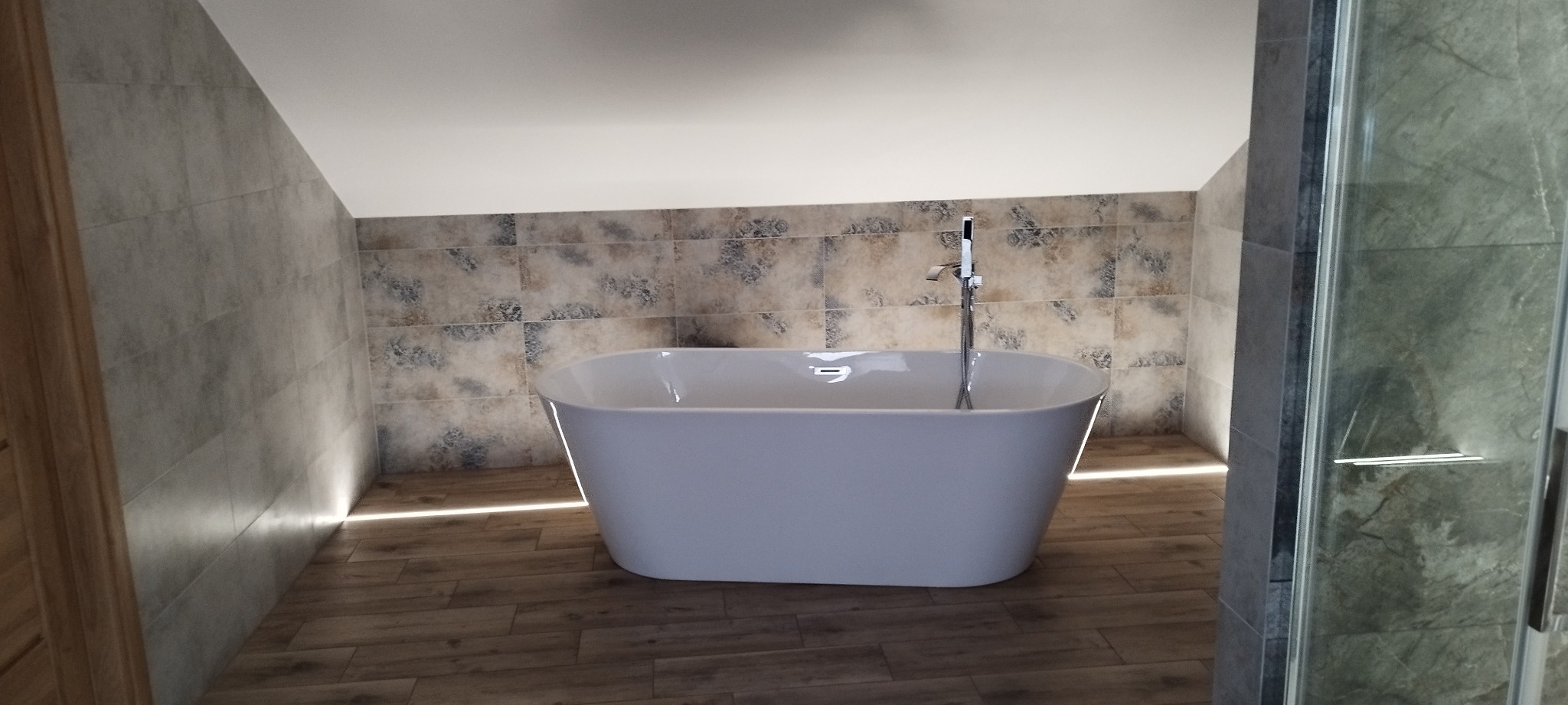 Nowoczesna łazienka na poddaszu z wolnostojącą białą wanną, drewnopodobnymi płytkami i podświetleniem LED przy podłodze. Kabina prysznicowa z marmurowymi płytkami.