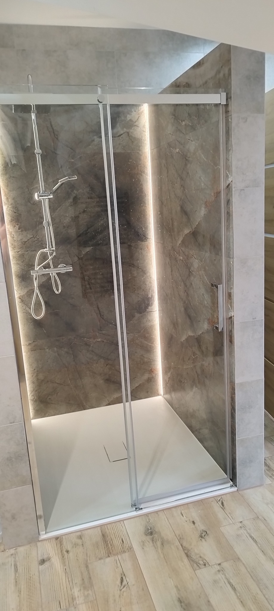 Nowoczesna kabina prysznicowa z przesuwnymi drzwiami i oświetleniem LED, wykończona płytkami imitującymi marmur. Biały brodzik i drewnopodobna podłoga.