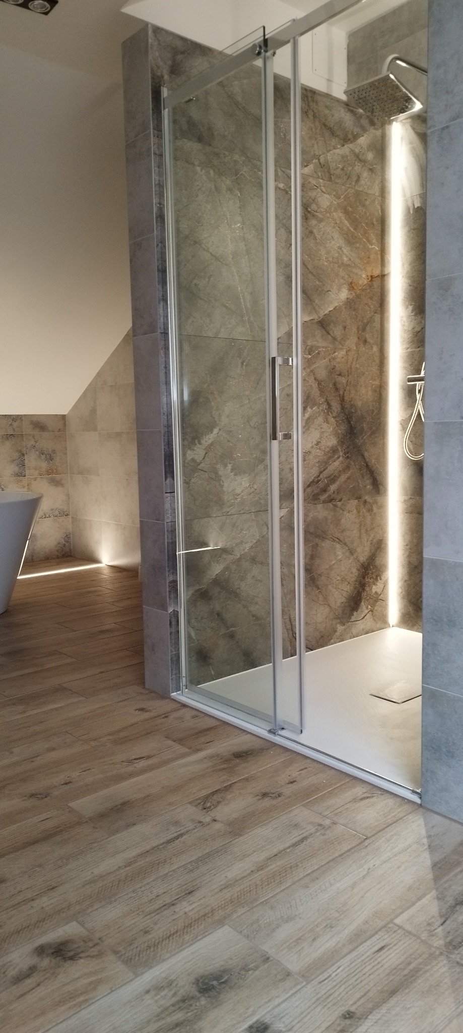 Nowoczesna kabina prysznicowa z marmurowymi płytkami i subtelnym podświetleniem LED, drewnopodobna podłoga,  elementy białej armatury.