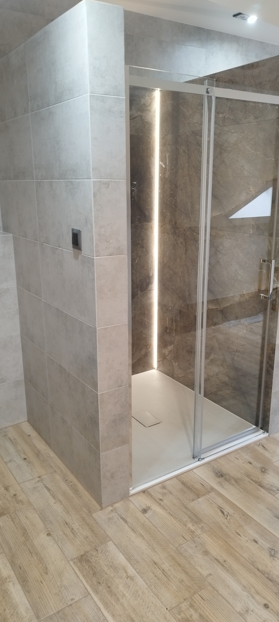 Wykończona kabina prysznicowa z oświetleniem LED i jasnymi płytkami imitującymi drewno na podłodze. Widoczne szare, kwadratowe płytki na ścianach.