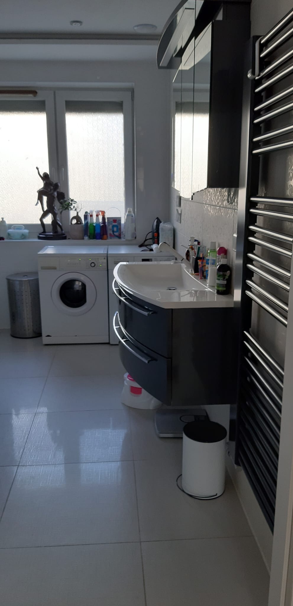 Nowoczesna łazienka z umywalką w zabudowie, pralką pod oknem i grzejnikiem drabinkowym. Jasne płytki podłogowe i dekoracje.