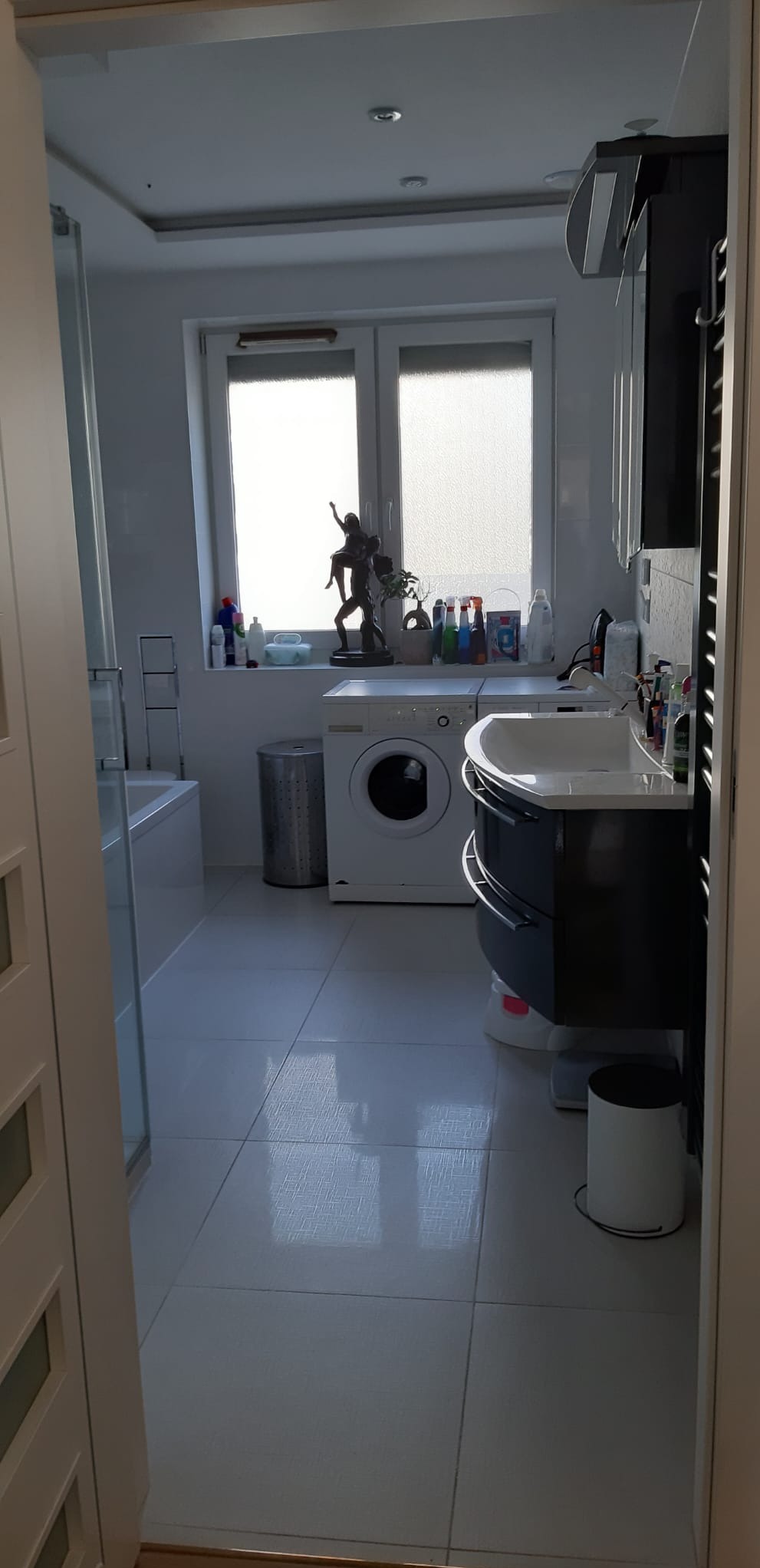 Jasna łazienka z oknem, pralką i umywalką z szafką. Na parapecie rzeźba i kosmetyki. Białe płytki podłogowe i drzwi. Widok z korytarza.