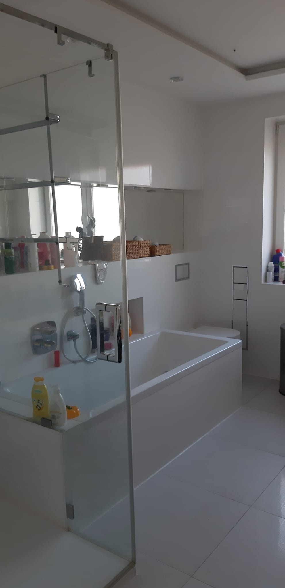 Jasna łazienka z wanną za szklaną ścianką, białe płytki podłogowe, minimalistyczne akcesoria i oświetlenie sufitowe.