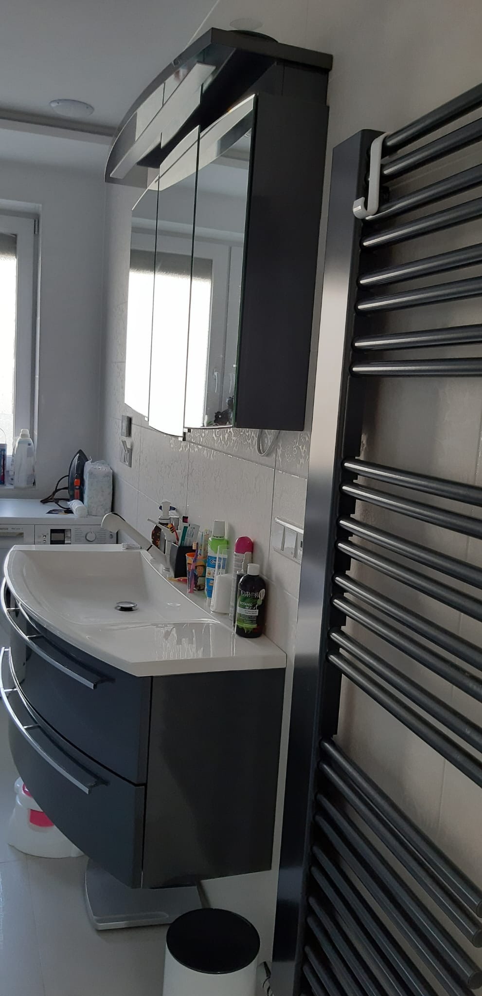 Nowoczesna łazienka z umywalką na szafce w kolorze grafitowym, lustrem w szafce i grzejnikiem drabinkowym. Na blacie kosmetyki i akcesoria łazienkowe.