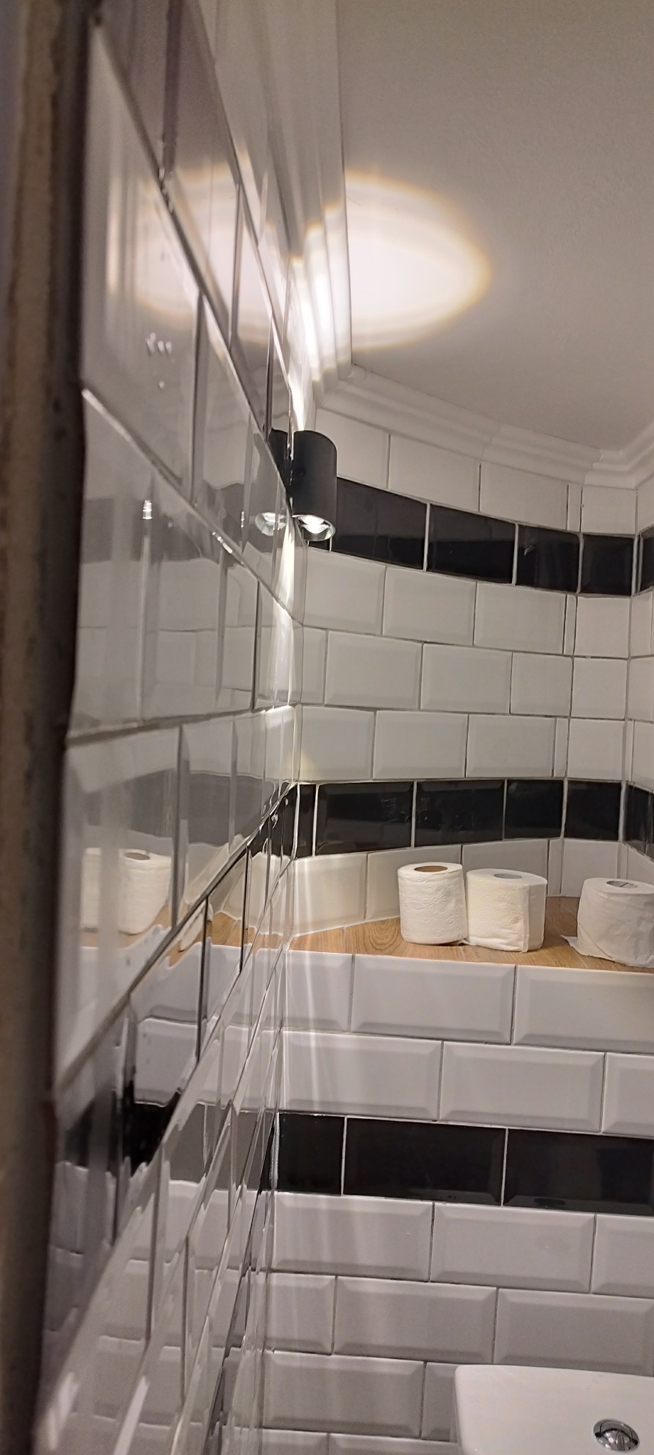 Białe, błyszczące płytki w stylu metro z czarnym pasem dekoracyjnym w wykończonej łazience. Na półce trzy rolki papieru toaletowego. Widoczna lampa ścienna.