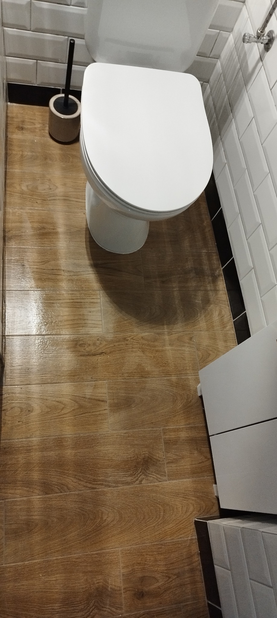 Biała toaleta na tle drewnopodobnych płytek podłogowych i białych, prostokątnych płytek ściennych z czarną fugą. Widoczny również element szafki pod umywalkę.