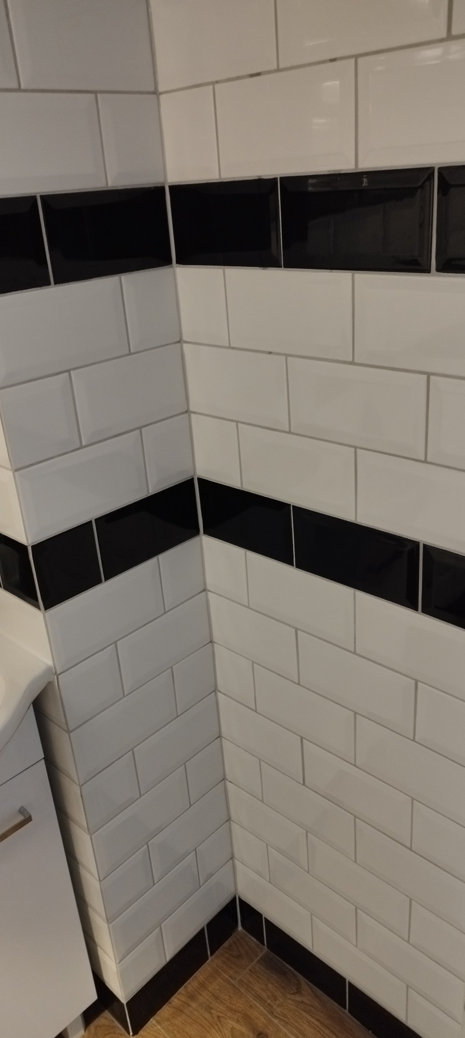 Wykończona ściana łazienki z białymi, prostokątnymi płytkami i poziomymi pasami czarnych płytek. Widoczny fragment szafki i podłogi.