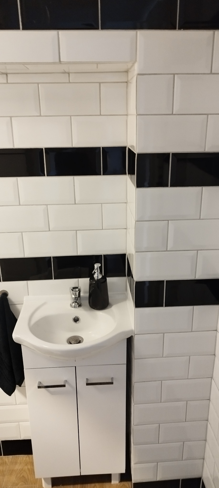 Biała umywalka z szafką w łazience z białymi płytkami, akcentami czarnych płytek i czarnym dozownikiem mydła. Widok na narożnik łazienki i drewnianą podłogę.