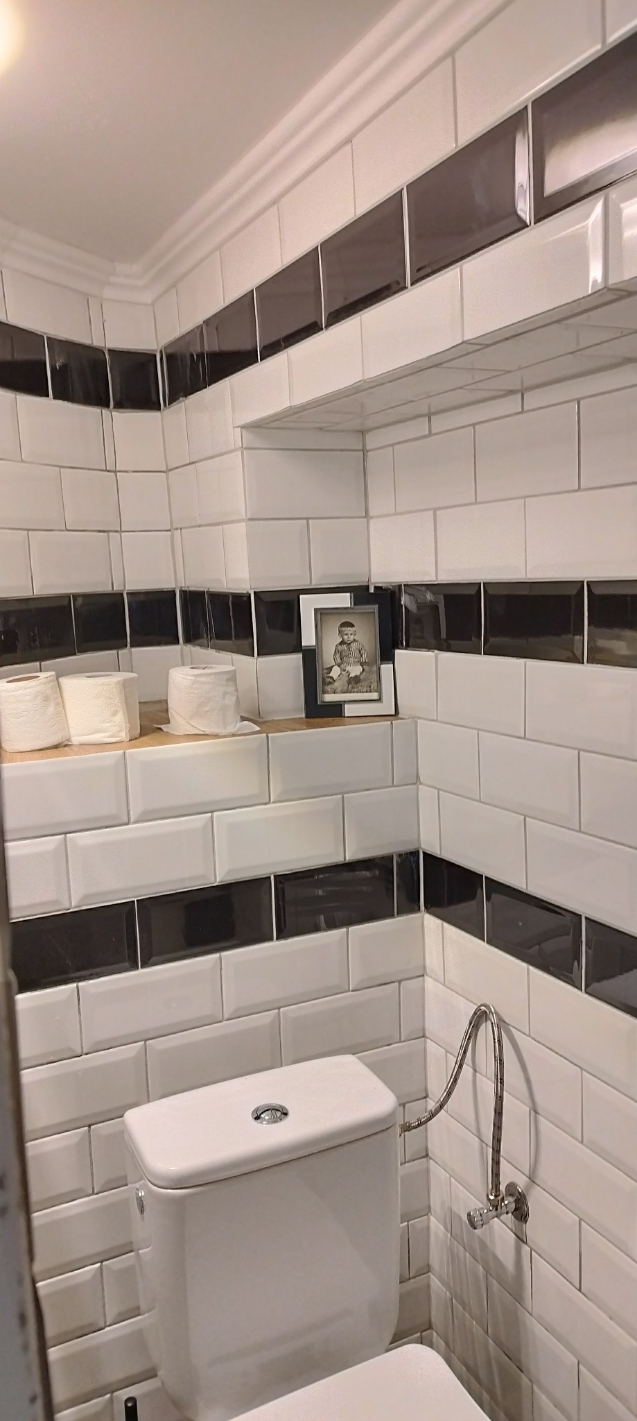 Biała toaleta z widocznymi białymi, prostokątnymi płytkami z czarnym pasem, drewnianą półką z papierem toaletowym i ramką ze zdjęciem.