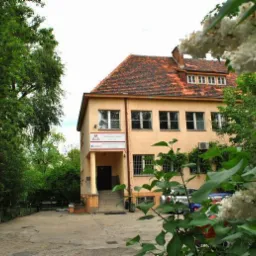 Dwupiętrowy budynek z pomarańczową dachówką, widoczny zza kwitnących bzów i zieleni drzew, z tabliczką informacyjną przy wejściu.