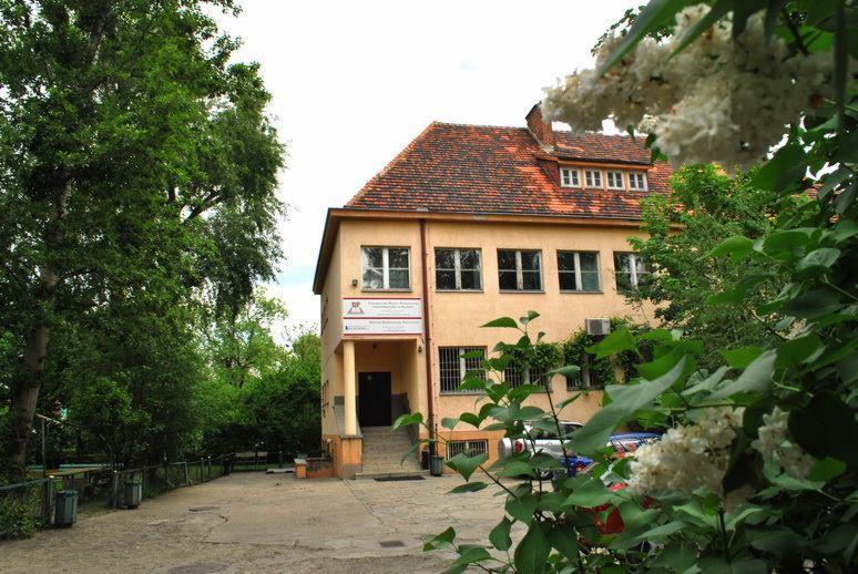 Dwupiętrowy budynek z pomarańczową dachówką, widoczny zza kwitnących bzów i zieleni drzew, z tabliczką informacyjną przy wejściu.