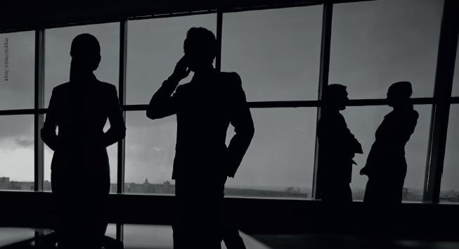 Sylwetki czterech osób w garniturach na tle okna z widokiem na miasto, mężczyzna w środku rozmawia przez telefon.